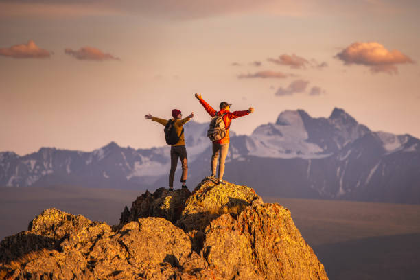 Two happy young tourists with backpacks are standing with open arms at mountain top and enjoys sunset view