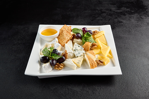 Cheese board with grapes and honey on a white plate against a black background.