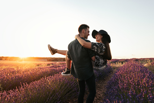 A young couple in love celebrates their anniversary with a romantic date in a lavender field at sunset. The guy picks up his girlfriend in his arms, and they walk among the flowers, looking at each other and smiling.
