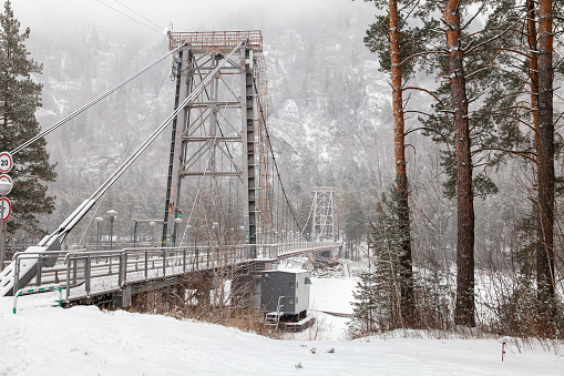 metal suspension bridge for cars and pedestrians in the Altai mountains in winter with snow and fog on the trees without people. Picturesque place winter landscape