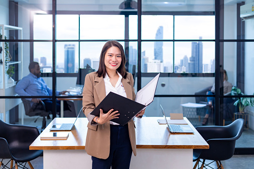 Portrait of Asian business CEO woman is standing in the office at the table with document file holder and showing the statistic chart showing report and skyscraper background