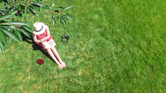 Young woman reading book in park near bike. Student girl relaxing outdoors sitting on grass with book and headphones, aerial drone view from above