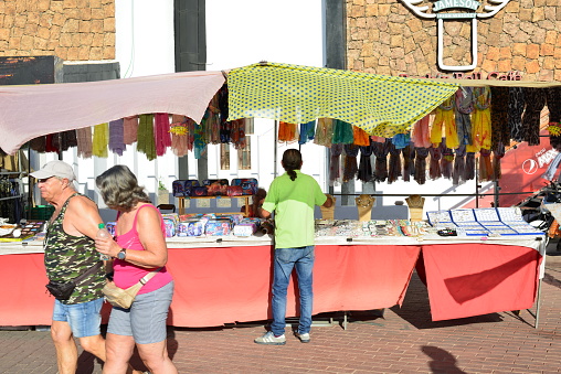 Gran Canaria, Canary Island of Spain - January 27, 2024: Market vendor stands at the front of his market stall of women's scarves and fantasy jewelry on a sunny winter day