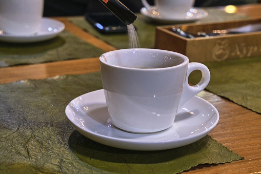 A hand pouring sugar from a packet into a cup of coffee.