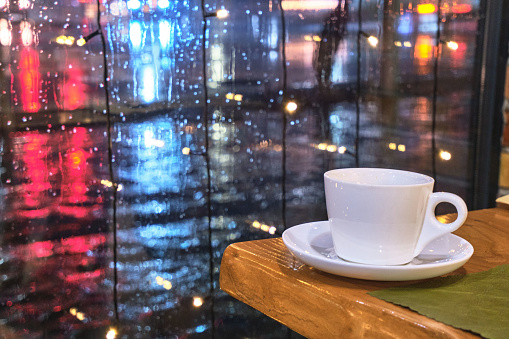 Coffee cup on the edge of a table near a window with rain outside.