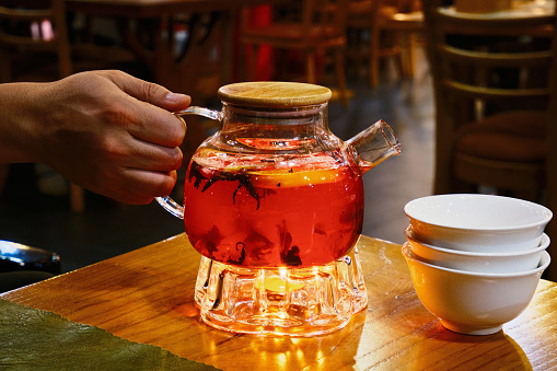 A hand holding a glass teapot with berry tea, and porcelain cups nearby.