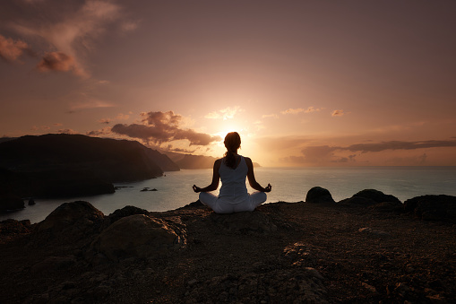 Back view of relaxed female athlete exercising Yoga on a mountain above the sea at sunset. Copy space.