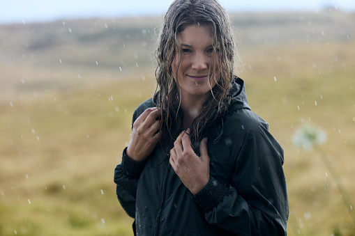 Portrait of smiling woman in raincoat enjoying during rainy day in nature and looking at camera. Copy space.