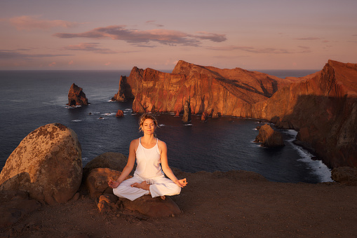 Relaxed athletic woman exercising Yoga in Lotus position during summer day on a cliff above the sea. Copy space.