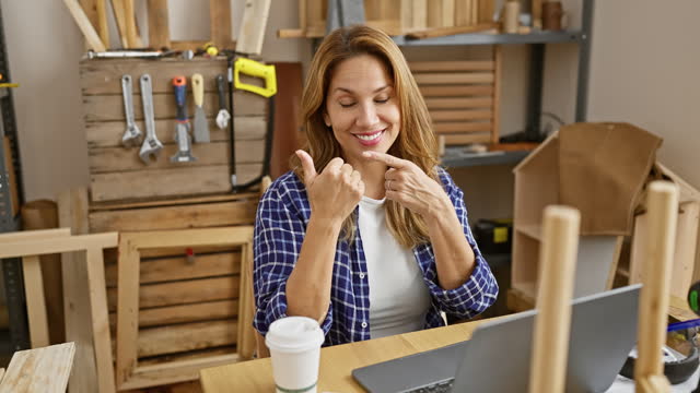 Confident young beautiful latin woman at carpentry, sitting at table, smiling and pointing behind with thumbs up, presenting her successful business.
