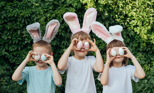 Smiling children wearing a headband with bunny ears found Easter eggs in the garden and brought them to their eyes. Siblings celebrate Easter. Religious holiday concept