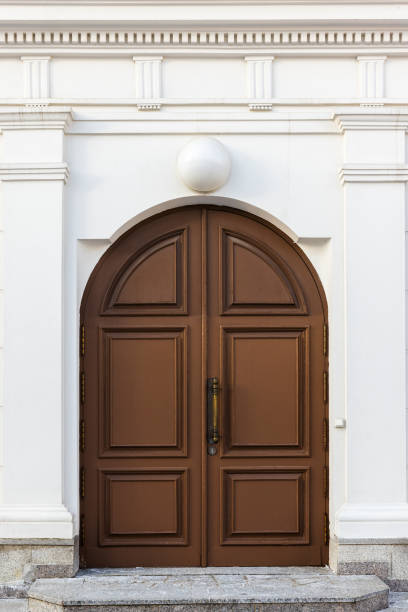 Brown wooden arched door in white wall with classic architecture decoration details, front view, background photo texture