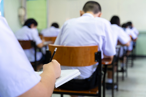 Soft focus.high school or university student holding a pencil writing on a paper answer sheet sitting on a chair lecturing on the final exam participating in an exam room or classroom students in uniform.