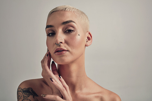 Model, portrait or tattoos in studio for beauty, cosmetics or make up in gen z aesthetic with piercings. Young woman, face or short blonde hair by nose ring in confidence or unique by gray background