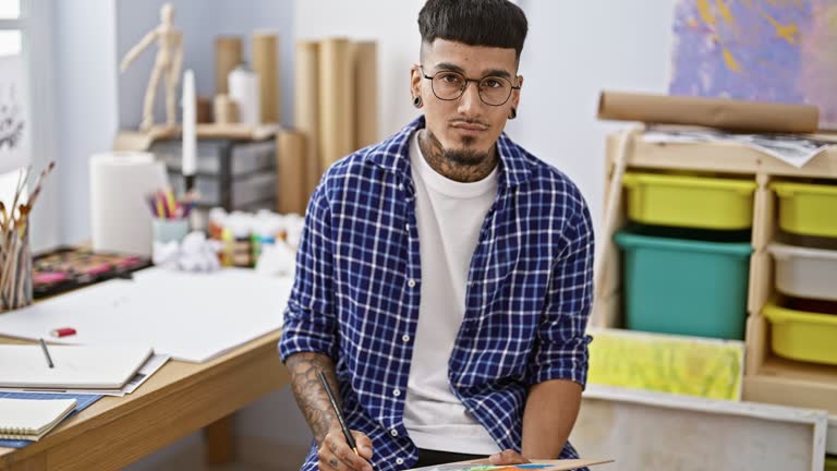 Handsome tattooed young latin artist intently wielding paintbrush and palette in his art studio - a portrait of concentration and creativity