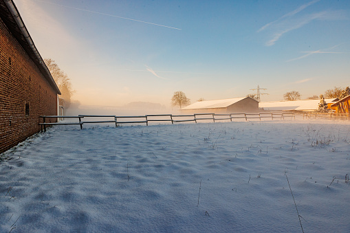 Agricultural plot covered in thick layer of snow after a heavy snowfall, wooden fence and farm in misty and blurry background, sunny sunrise in Beek, South Limburg, Netherlands