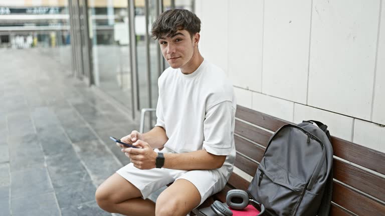 Chilled young hispanic guy, a confident university student, gives thumb up gesture while sitting on a bench, engrossed in his smartphone, amidst the casual urban college lifestyle.