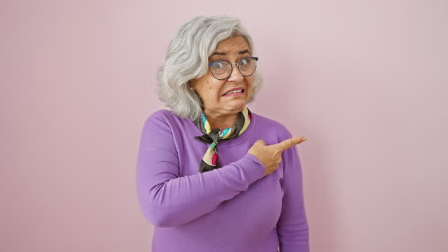 Worried grey-haired woman in glasses, nervously pointing aside with surprised expression, showing concern over shocking mistake on isolated pink background