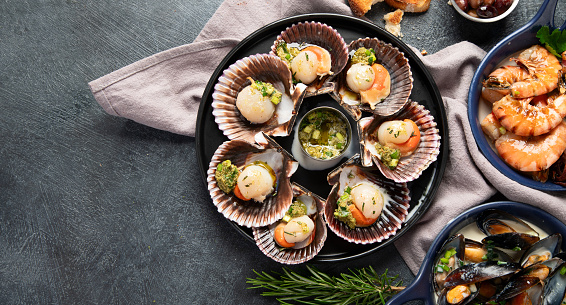 Baked scallops in seashells with sauce and spices on a black stone plate on a dark background. Seafood. Top view. Copy space.