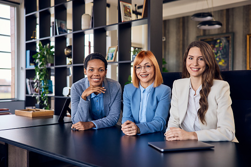 Three smiling females, sitting at the office, posing for the camera, dressed elegantly.