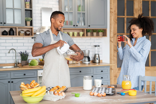 Couple cooking together, having fun time in the kitchen. Multiracial man mixing eggs in a bowl, while his girlfriend taking picture of him with her mobile phone.