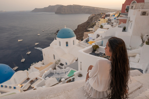 Asian female tourist on a vacation in Santorini, standing on a balcony and enjoying the view on sunset