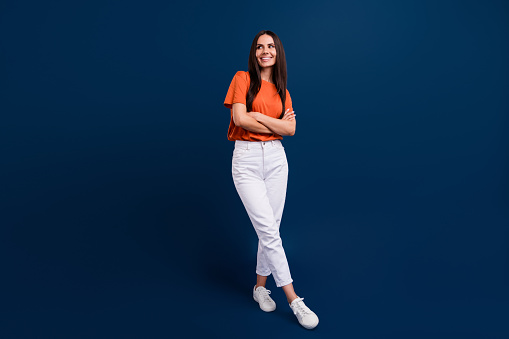 Full length photo of shiny dreamy woman dressed orange t-shirt arms folded looking empty space isolated dark blue color background.