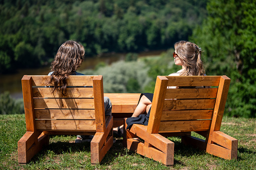 Two girls are sitting on large wooden chairs in a park on a high mountain in the forest. Woman's talk. Sunny summer day. Back view