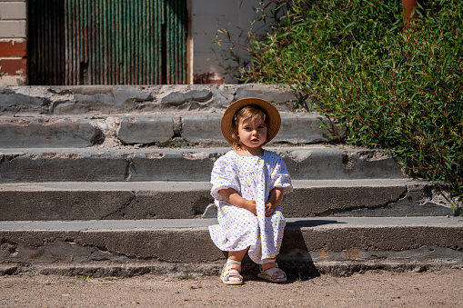 A cute little girl in a dress and hat sitsing on old concrete steps. Sunny summer day.