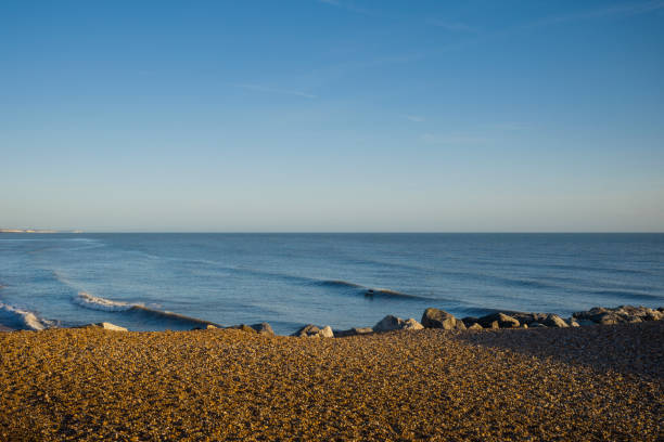 A pebble beach with a sea horizon on a sunny day, and a lone and unrecognisable surfer in the mid-distance.