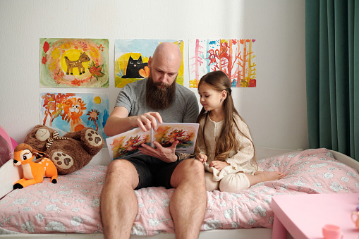 Bearded man with open book of comics with pictures sitting on bed next to his adorable little daughter and reading her new stories