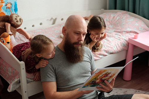 Bearded father holding open book of comics while sitting on the floor by bed with his two adorable daughters looking at pictures