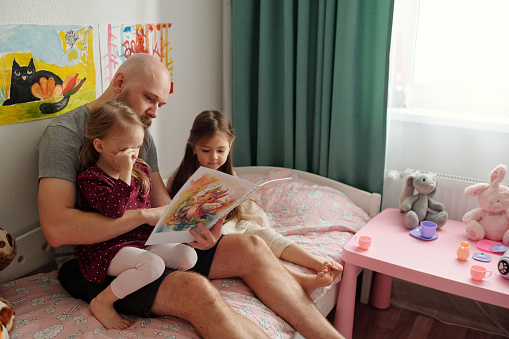 Bearded man with bald head reading book of comics with pictures to his cute little daughters while sitting on bed between them
