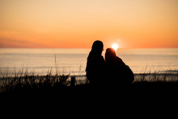 Silhouette of two female friends enjoying a conversation while sitting hugging and watching the sunset on a sea beach. Back view
