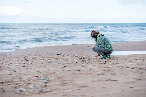 Alone teenager girl in green hoodie stacking stones on the Baltics seashore.