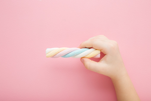 Little girl hand fingers holding colorful swirl soft marshmallow candy on light pink table background. Pastel color. Closeup. Children sweet snack. Top down view.