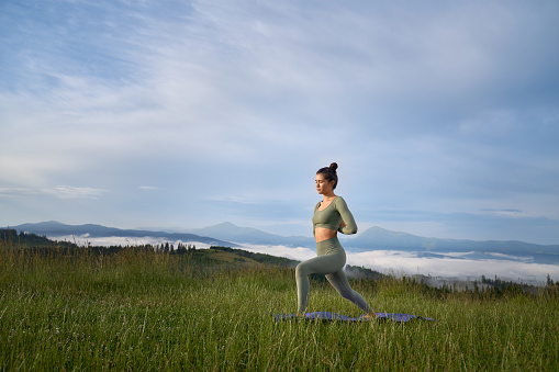 Attractive woman in sport clothes having outdoors workout during warm summer day. Relaxed young lady enjoying yoga practice among mountains.