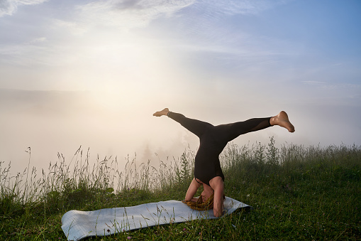 Active woman with dreadlocks standing on head and splitting legs with amazing view on summer sunrise. Young lady in black sport clothes enjoying morning workout outdoors.