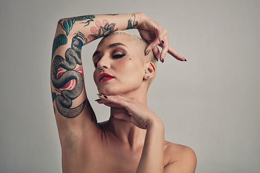 Tattoo, piercing and face of woman with beauty in grey background of studio with confidence. Unique, girl and person with edgy or punk aesthetic with makeup, skincare and pride for ink on body