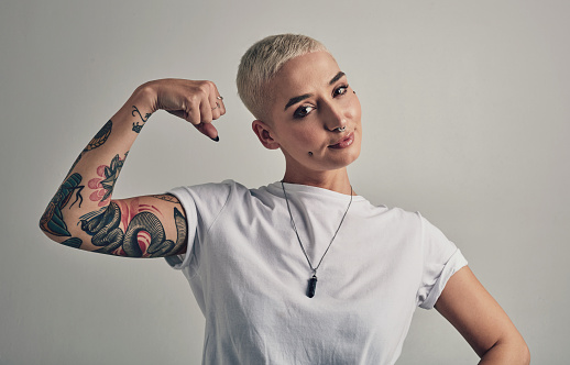 Power, flex and portrait of woman with tattoo fashion, creative culture and confidence in studio. Artistic girl with muscle, identity and trendy style with hipster clothes, pride and grey background.