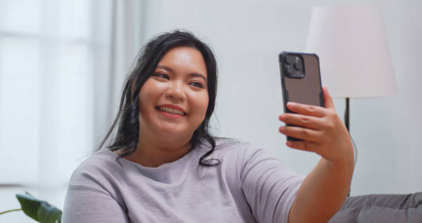 istock A chubby Asian woman using smart phone to take selfie while sitting on sofa at home. 1972577817