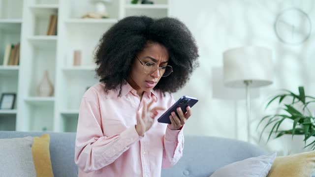 Angry african american female complains about poor performance of program on smartphone sitting on sofa at home