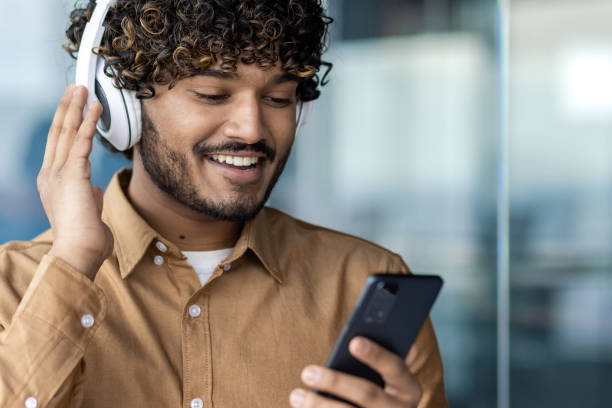 a young man with curly hair smiles as he listens to music through his white headphones, holding a smartphone in a brightly lit modern setting, expressing pure enjoyment. - brightly lit audio imagens e fotografias de stock