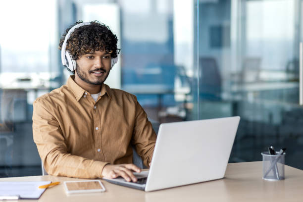 istock Young indian programmer working inside office with laptop, man in headphones coding new software, businessman developer satisfied with achieved results smiling 1972663910