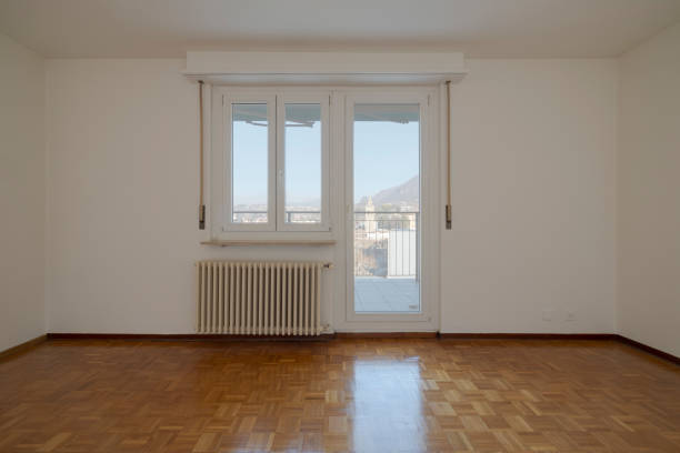 Empty living room with a bright window looking out at the blue sky. An old radiator hangs on the white walls. No one inside