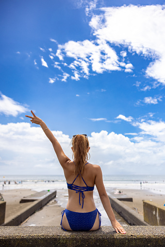 Rear view of woman in bikini sitting at beach - one hand raised
