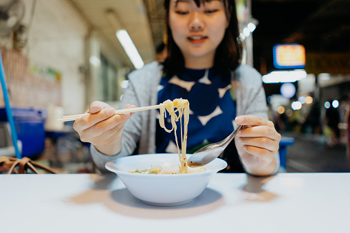 An Asian female tourist enjoying street food Tom Yum noodle soup at a night market in Bangkok
