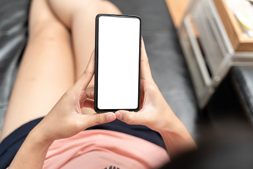 Young woman or teen girl hands holding cell phone touching finger mockup white blank display, empty screen for social media app ad at home. Mobile application tech concept, over shoulder closeup view.