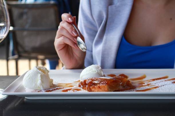 Close-up of young woman eating dessert in restaurant at daytime. Female's hands holding fork with piece of cake and knife over plate of cherry pie served with scoop of ice cream.