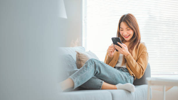 istock Happy young asian woman browsing surfing wireless internet on mobile smartphone while sitting a couch in living room at home, Shopping online via website 1972736475